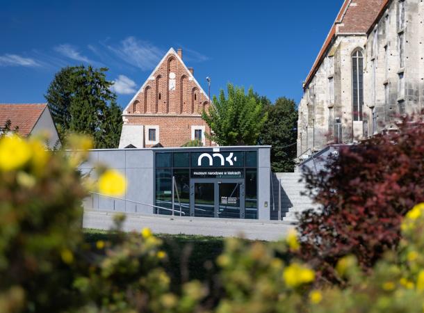 Fotografia nowoczesnego sklepu z logo mk przed zabytkowym kościołem, rozmyte żółte kwiaty