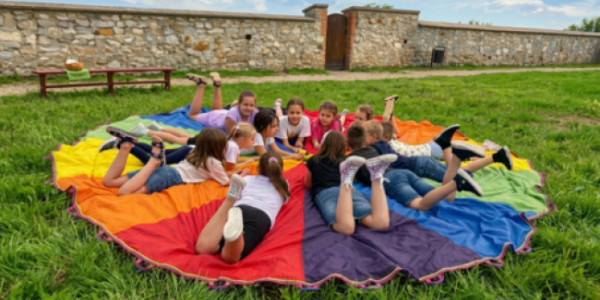 Fotografia: grupa dzieci leżąca w kręgu na kolorowym, tęczowym płótnie na trawie