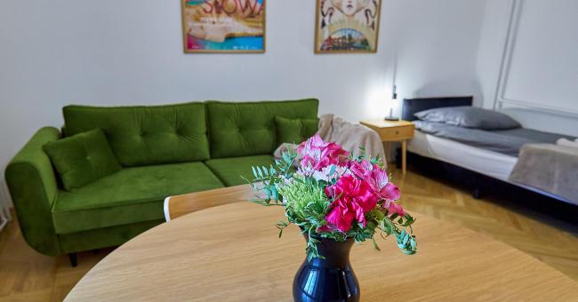 Fotografia wnętrza: zielona kanapa, stół z czarnym wazonem i różowymi kwiatami