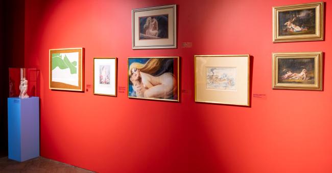 Czerwona ściana galerii z oprawionymi obrazami i fotografią aktu; niebieski piedestał z rzeźbami