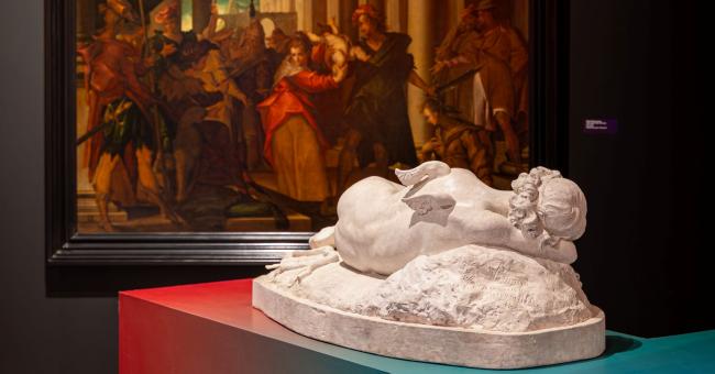 Fotografia wystawy: obraz na ścianie i biała rzeźba leżącej postaci na kolorowych postumentach