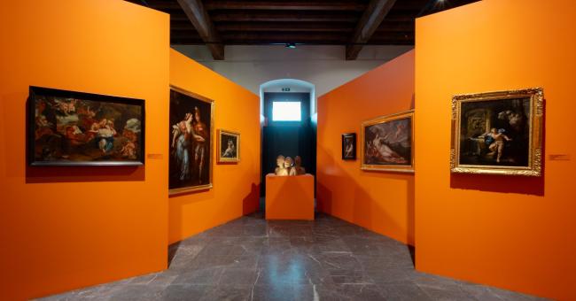 Fotografia wnętrza galerii z pomarańczowymi ścianami, obrazami i rzeźbą na postumencie