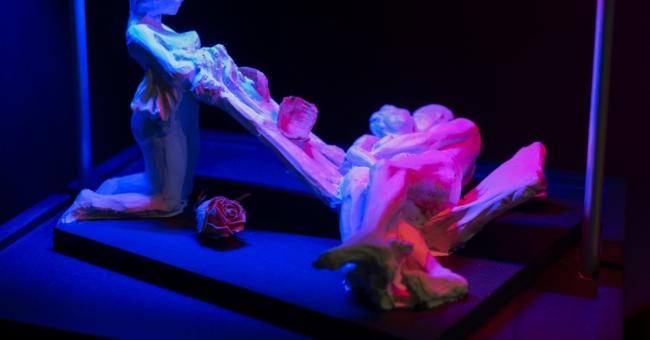 Fotografia białych rzeźb: stojąca postać ciągnie tkaninę z leżącej; niebiesko-różowe światło