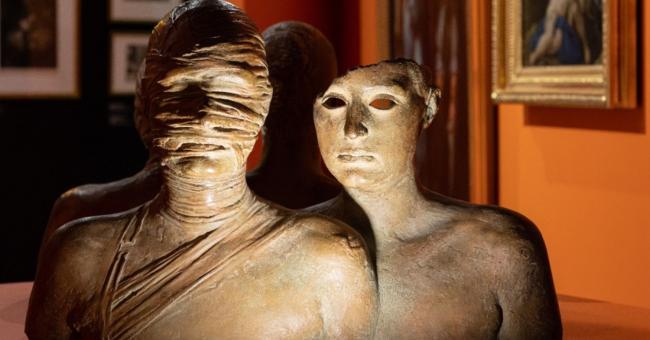 Fotografia rzeźby: dwa popiersia — obandażowana twarz i koci profil, pomarańczowe tło.