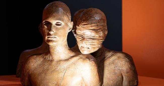 Fotografia dwóch brązowych torsów rzeźb; jeden z twarzą zabandażowaną, pomarańczowe tło