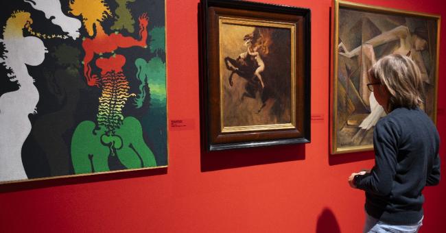 Fotografia: osoba ogląda trzy obrazy na czerwonej ścianie galerii