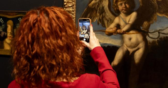 Fotografia kobiety z rudymi włosami fotografującej obraz anioła z dzieckiem w złotej ramie