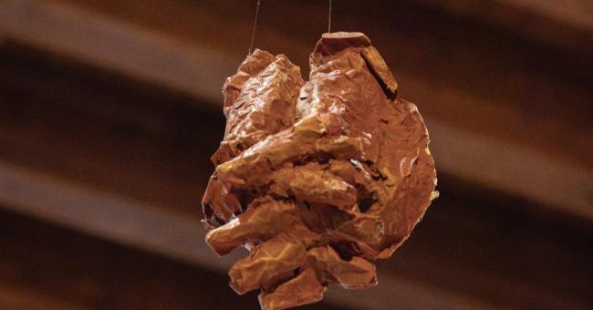 Fotografia wiszącej glinianej rzeźby przypominającej dłoń, brązowa, z drewnianym sufitem w tle.