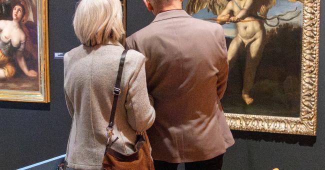 Fotografia: starsza para ogląda obrazy w galerii, złote ramy na ciemnej ścianie