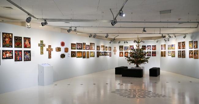 Fotografia wnętrza galerii z religijnymi obrazami i krzyżami, choinka w centrum