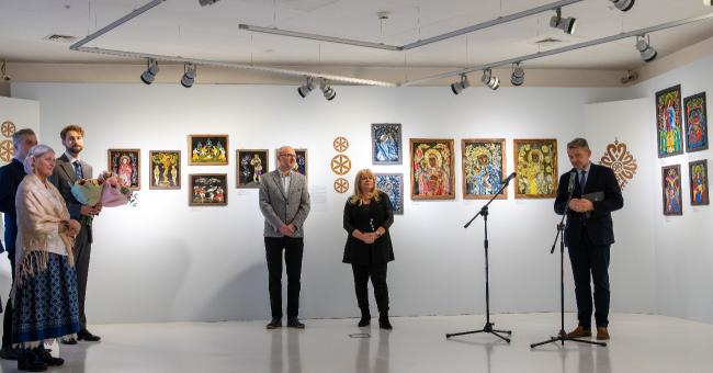 Wernisaż w galerii: grupa ludzi i trzech przemawiających przy mikrofonach, kolorowe obrazy namalowane na szkle na ścianach