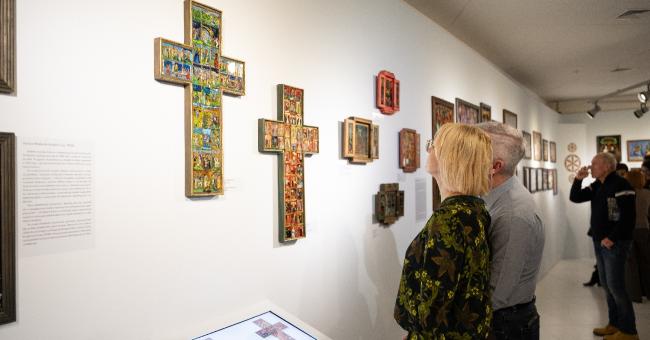 Dwie osoby w galerii oglądają kolorowe, ozdobne krzyże na białej ścianie