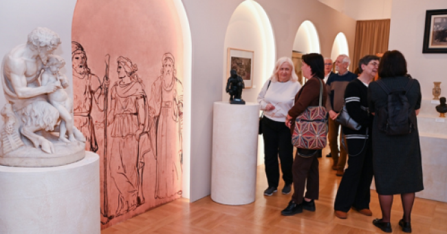 Fotografia sali wystawowej z białymi rzeźbami i grupą zwiedzających przy różowej ścianie