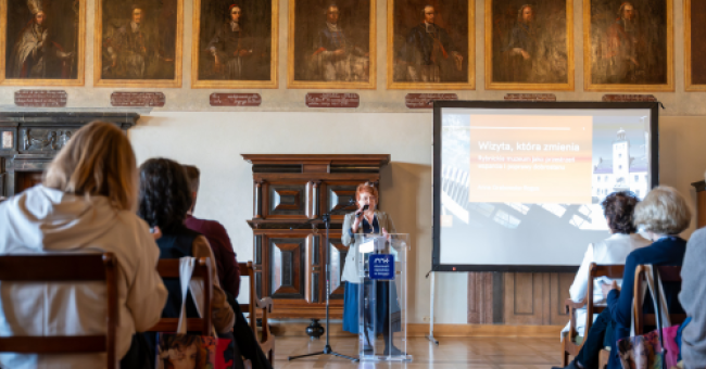 Fotografia: osoba przemawia przy mikrofonie w zabytkowej sali, portrety na ścianie, publiczność