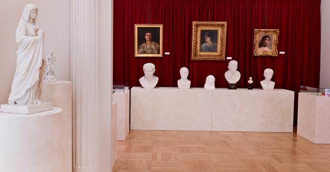 Fotografia wnętrza galerii: białe rzeźby i popiersia na tle czerwonej kotary z portretami.