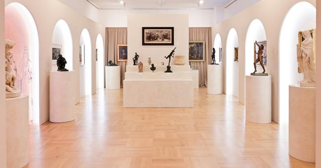 Fotografia jasnej galerii z rzeźbami na postumentach, łukowymi niszami i pastelowym oświetleniem