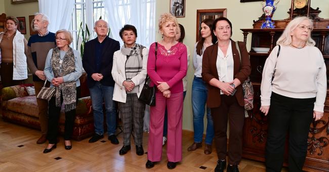 Fotografia: grupa dorosłych stoi w umeblowanym salonie, kobieta w różowym garniturze pośrodku