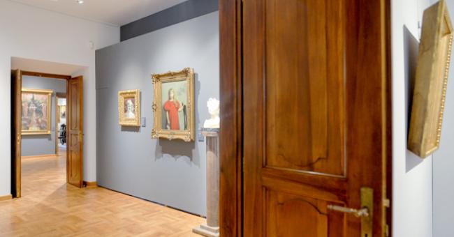 Fotografia wnętrza galerii: otwarte drewniane drzwi, obrazy w złotych ramach i popiersie