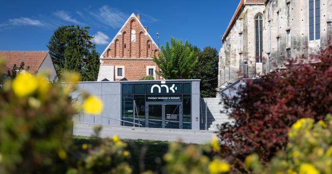Czarny kiosk z białym logo przed zabytkowymi budynkami, rozmyte żółte kwiaty