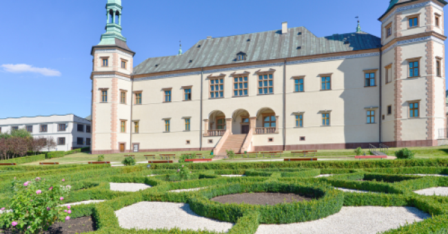 Fotografia zabytkowego pałacu z symetrycznym, przyciętym ogrodem i jasnym niebem