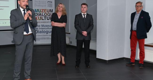 Fotografia: cztery osoby w galerii, mężczyzna przemawia do mikrofonu