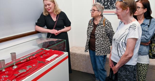 Fotografia: przewodniczka opowiada grupie kobiet przy czerwonej gablocie muzealnej.