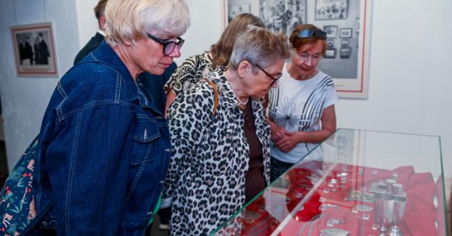 Fotografia: trzy starsze kobiety oglądają eksponaty w szklanej gablocie na czerwonym tle