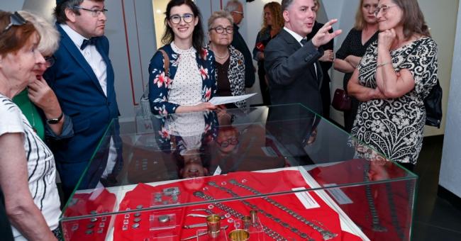 Grupa osób przy szklanej gablocie z czerwonym strojem i odznaczeniami