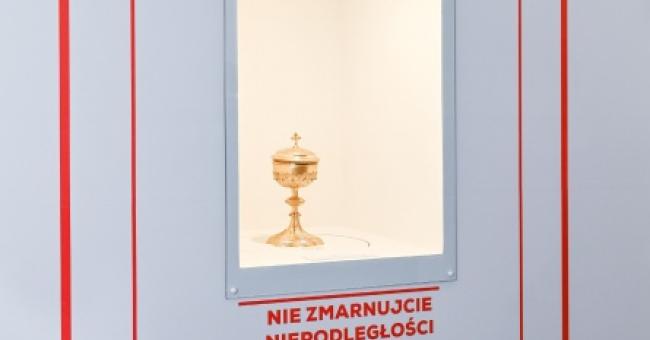 Plakat wystawowy z czerwonym ramowaniem i podświetlonym złotym kielichem w niszy