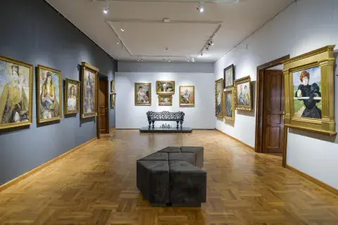 Fotografia sali galerii z obrazami w złotych ramach, dwoma siedziskami i drewnianym parkietem
