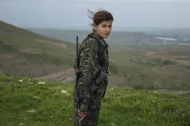 w centrum zdjęcia kobieta ubrana w mundur  kaki z przewieszonym karabinem na ramieniu na tle gór pokrytych niskopienną roślinnością, włosy kobiety rozwiewa wiatr.